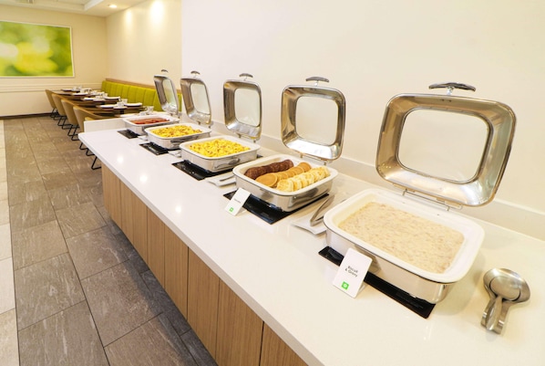 Una barra de buffet con diferentes platos en recipientes metálicos calientes, un área de estar con sillas y un banco acolchado verde.