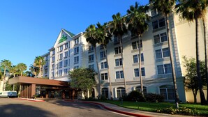A multi-story hotel with a prominent green logo on the facade, a covered entrance, and palm trees in the surrounding area. - Wyndham Garden Mcallen at La Plaza Mall (McAllen)