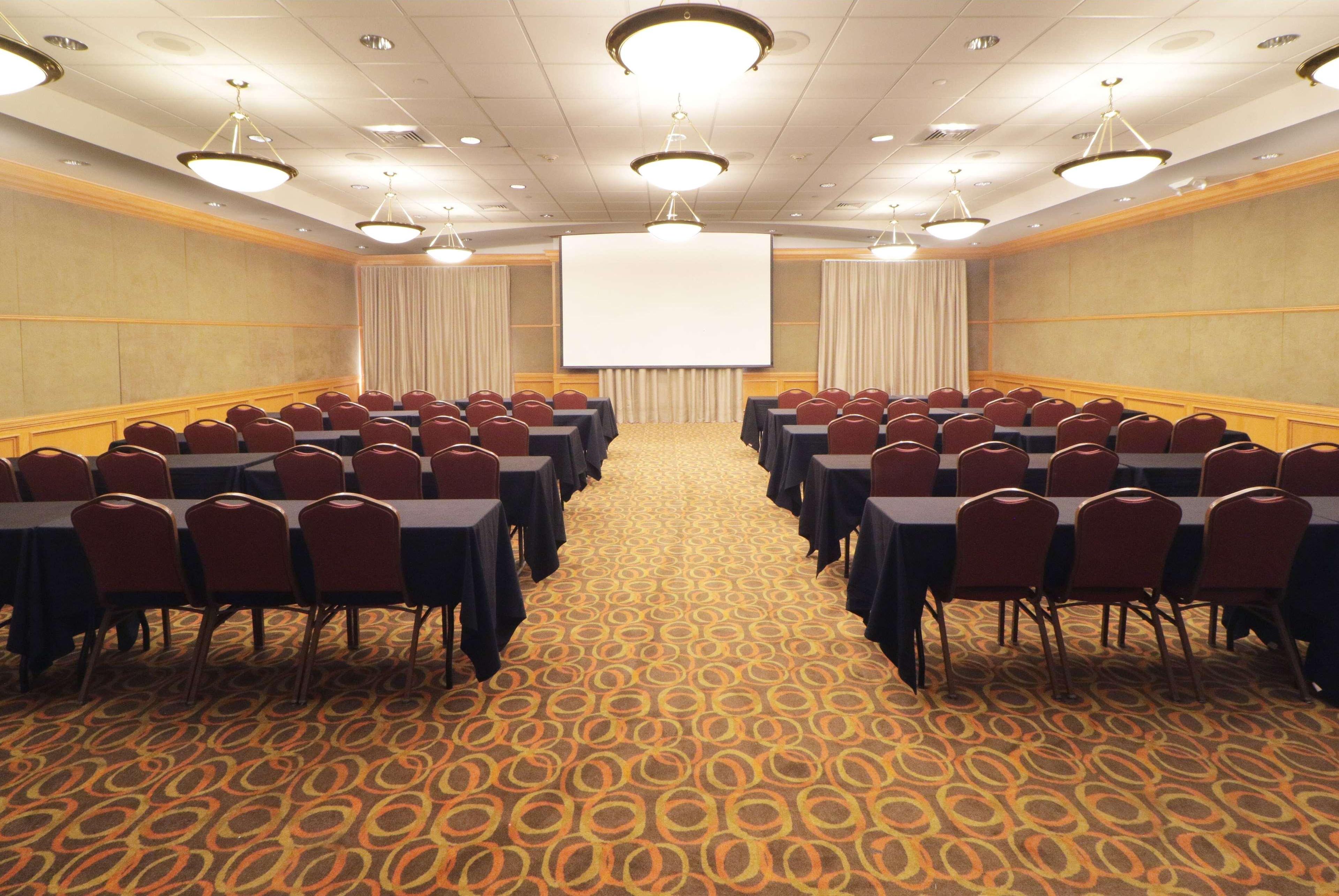 A conference room with rows of tables and chairs, a large screen at the front, and a patterned carpet.