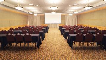 A conference room with rows of tables and chairs, a large screen at the front, and a patterned carpet.
