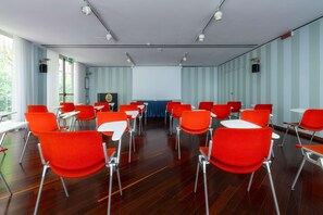 Een vergaderruimte met rode stoelen en tafels, grote ramen en een scherm voor een beamer.