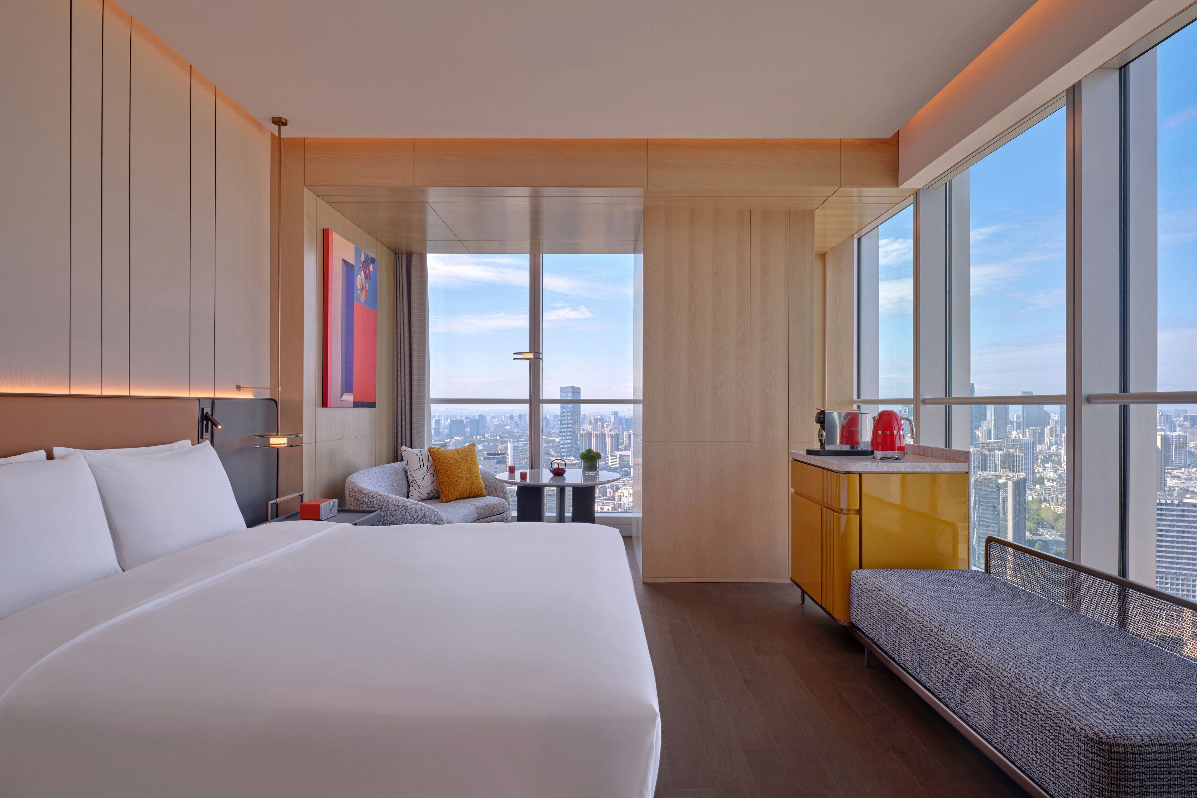 Ein modernes Hotelzimmer mit einem großen Bett, einer Sitzecke, einem kleinen Tisch und Blick auf die Stadt durch große Fenster.