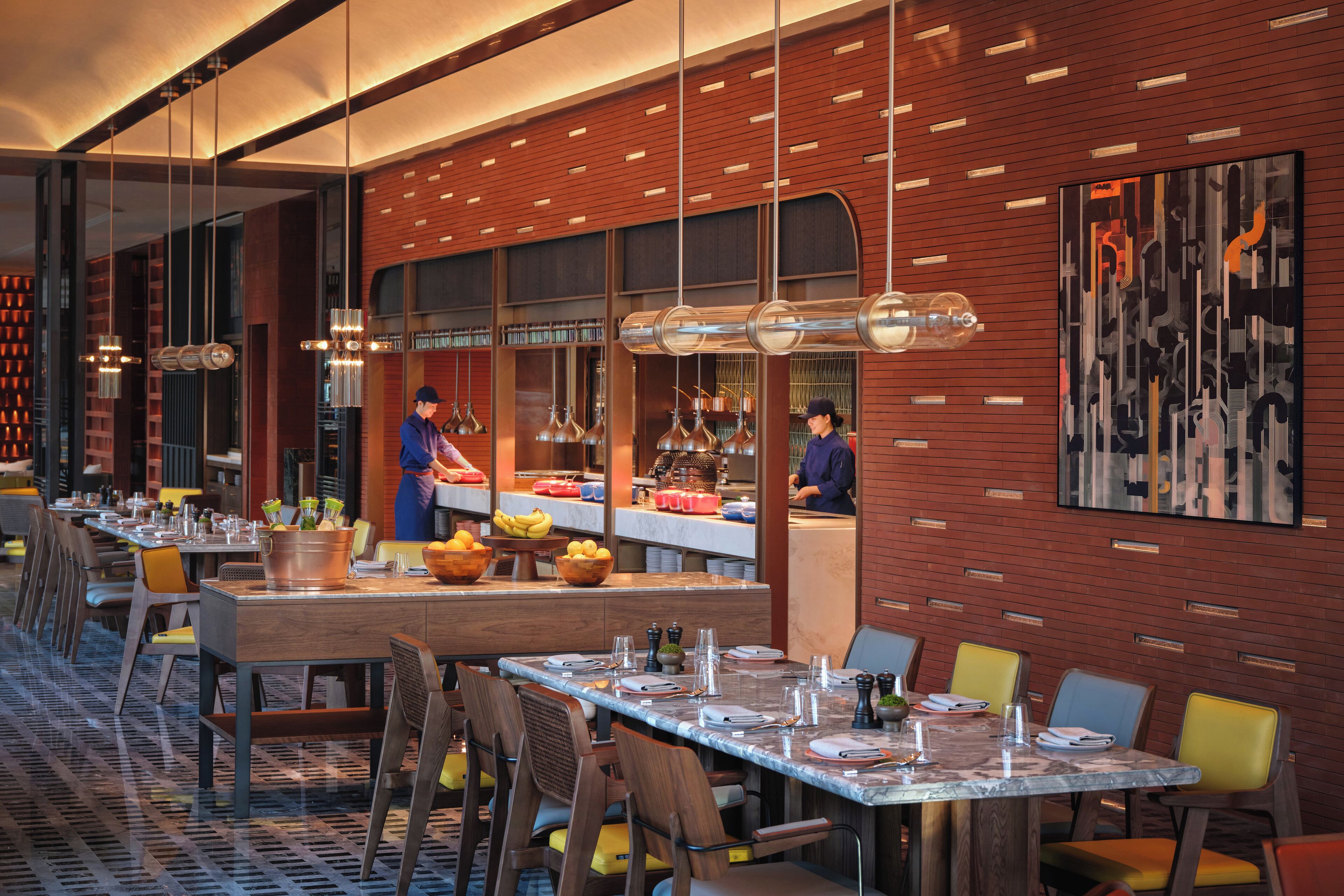 A modern restaurant with marble tables, yellow and brown chairs, and a brick wall with a large abstract painting.