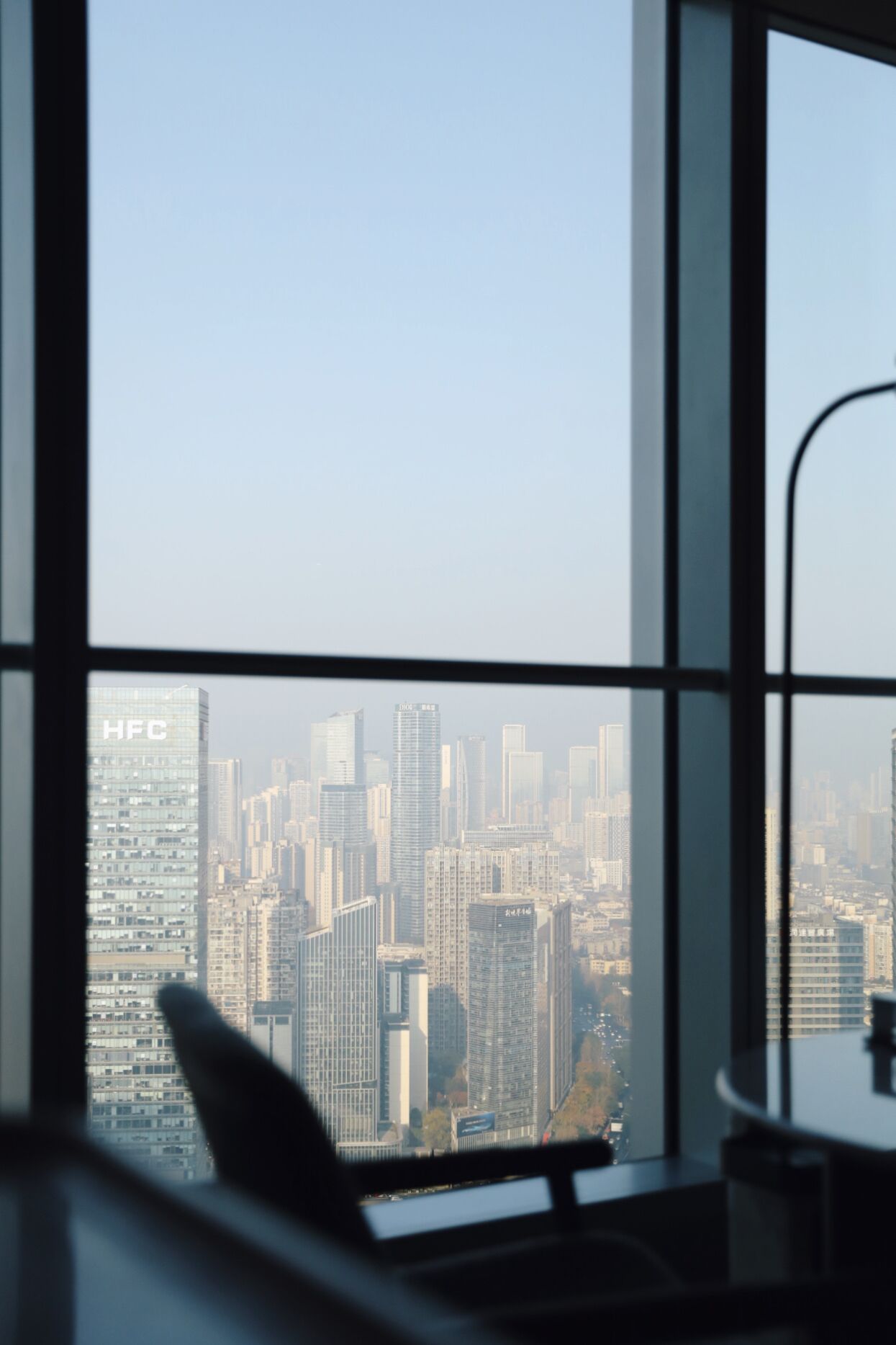 Blick auf eine Stadtansicht durch ein Fenster, im Vordergrund ein Schreibtisch und ein Stuhl.
