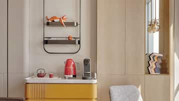 A modern kitchen with a yellow cabinet, a red kettle, and a coffee maker. There is a chair and a table with a book and a mug.