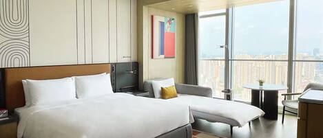 A hotel room with a large bed, a desk, a chair, a small table, and a view of the city.