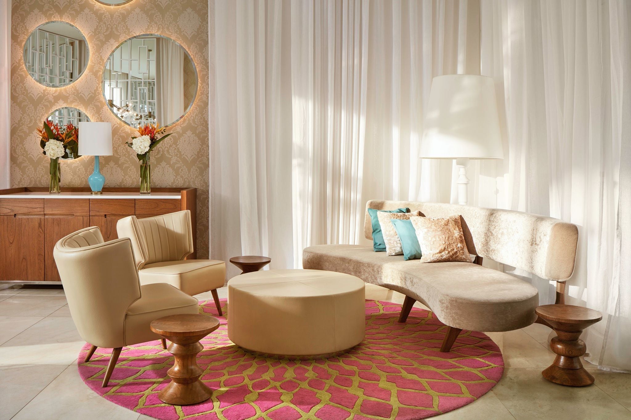 A modern living room with a beige sofa, a round ottoman, and a wooden coffee table. There are two lamps on side tables, and a large mirror on the wall.