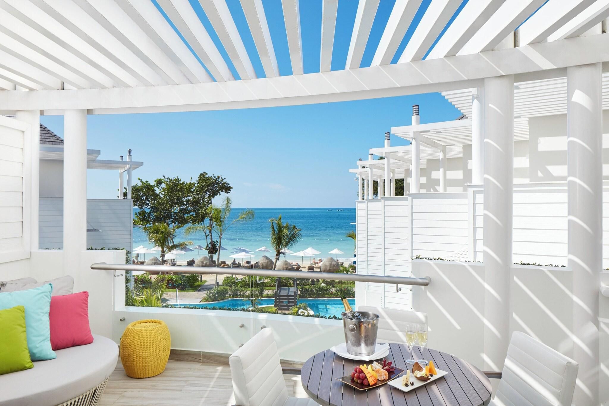 Un balcon avec vue sur l’océan, une piscine et une table avec une boisson et des fruits.