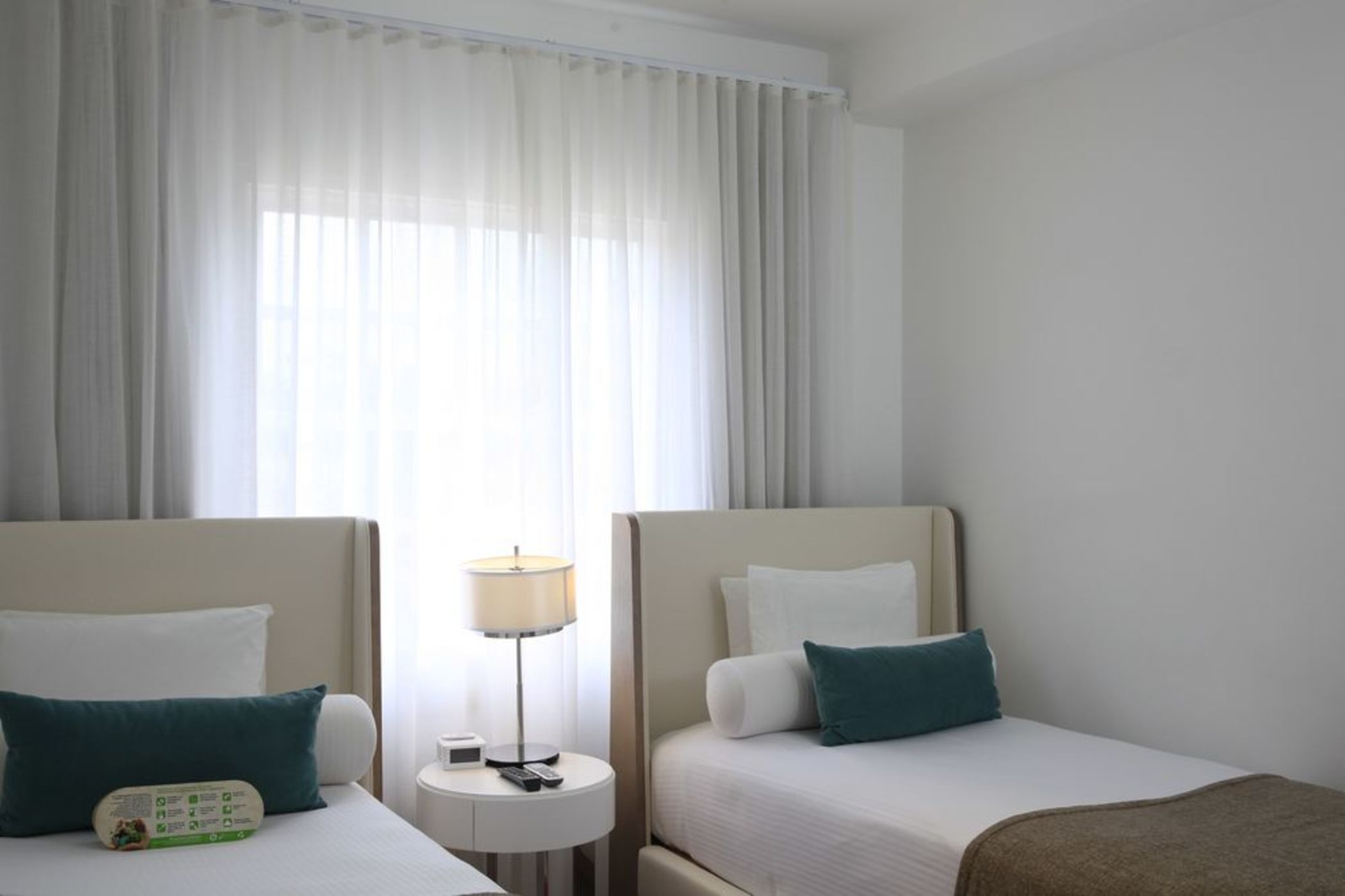 A hotel room with two beds, a nightstand with a lamp, and a window with curtains.