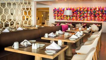 A modern restaurant with long communal tables, cushioned seating, and a vibrant, colorful wall decoration.