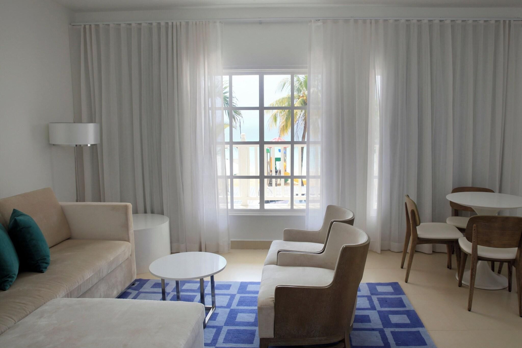 A modern hotel room with a sofa, a round coffee table, a dining set, and a large window with curtains.