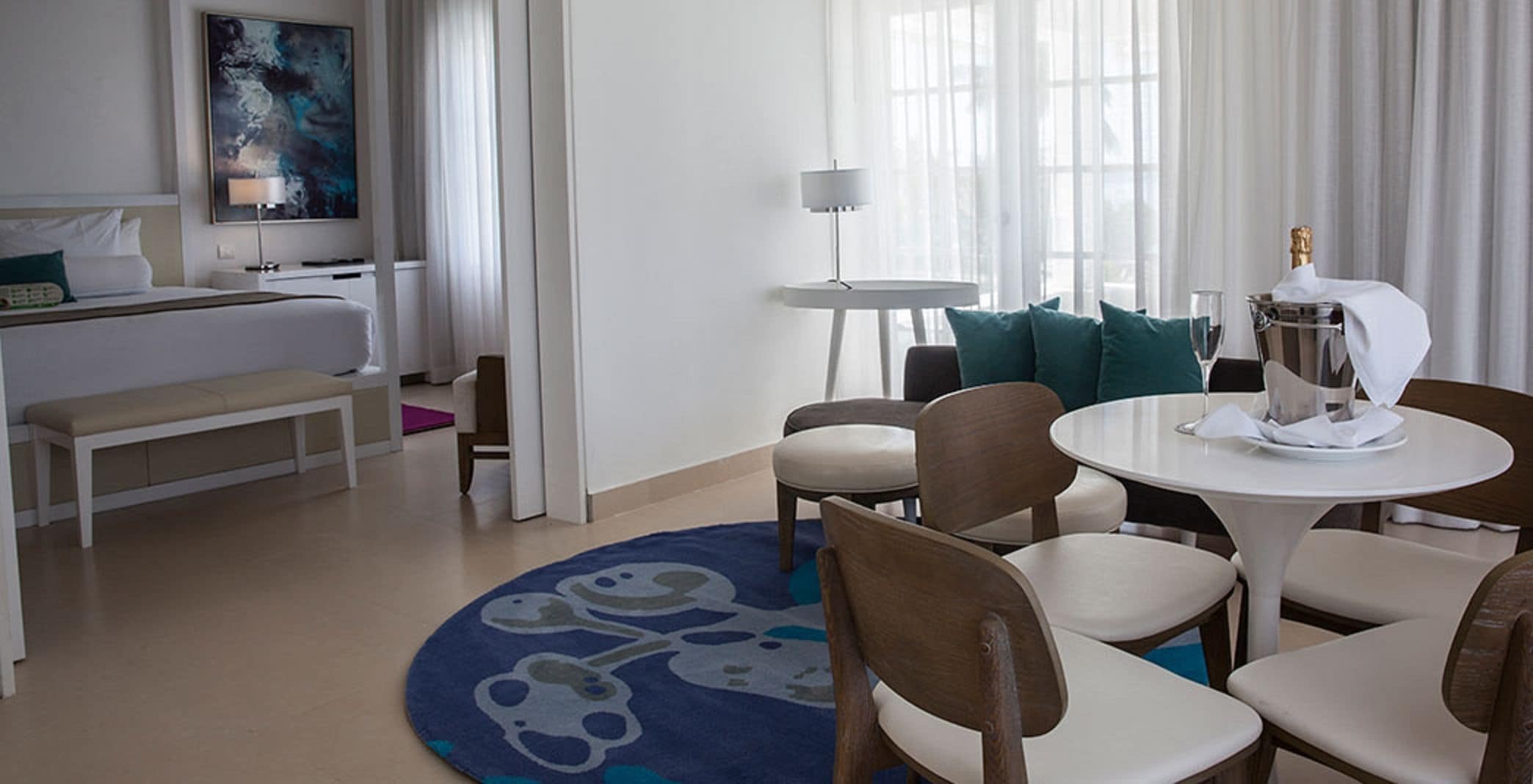 Una habitación de hotel con cama, escritorio, mesa de comedor con sillas, una mesita redonda con lámpara y un ventanal con cortinas.