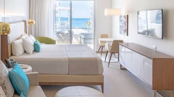 A hotel room with a bed, a sofa, a desk, and a TV. There is a balcony with a view of the sea.
