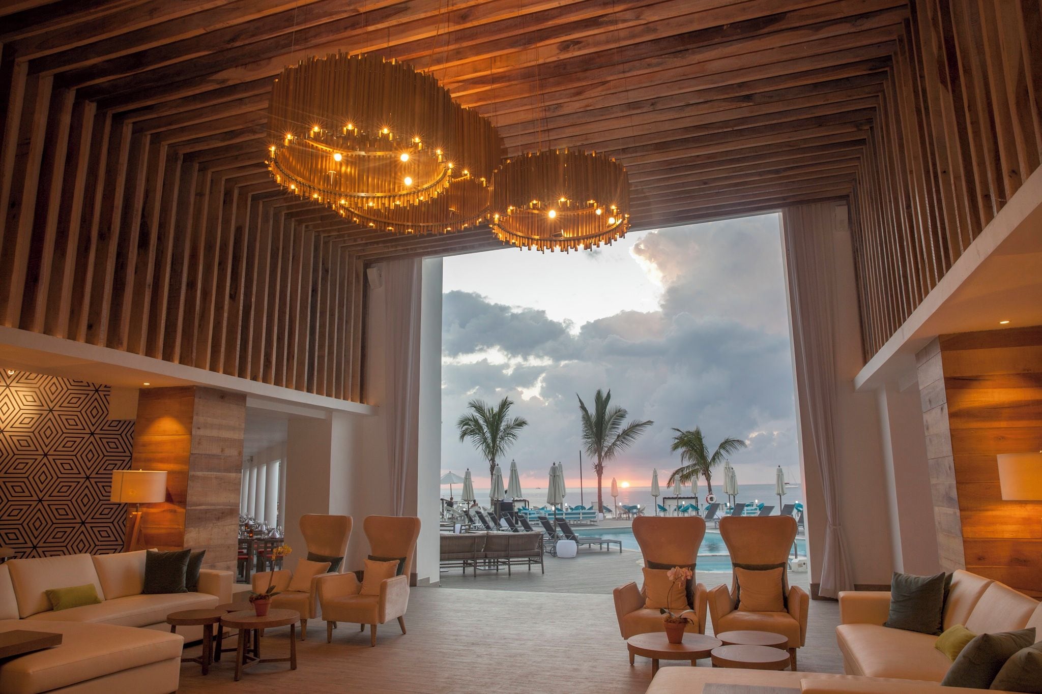 A spacious lobby with a large window overlooking a beach, furnished with sofas and armchairs, and featuring a wooden ceiling and patterned wall.
