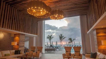 A spacious lobby with a large window overlooking a beach, furnished with sofas and armchairs, and featuring a wooden ceiling and patterned wall.