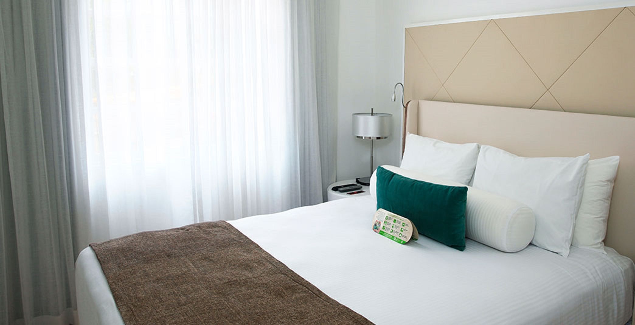 Quarto de hotel com cama, mesa de cabeceira, abajur e janela com cortinas.