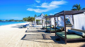 Un complexe hôtelier en bord de mer, doté de bungalows sur pilotis, offrant une vue sur un ciel bleu azur et donnant accès à une plage de sable fin.
