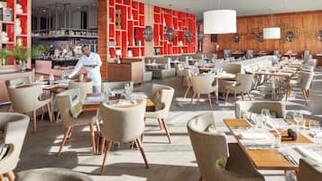 Restaurante moderno com amplo espaço para refeições, várias mesas e um bar, onde um chef está a servir.