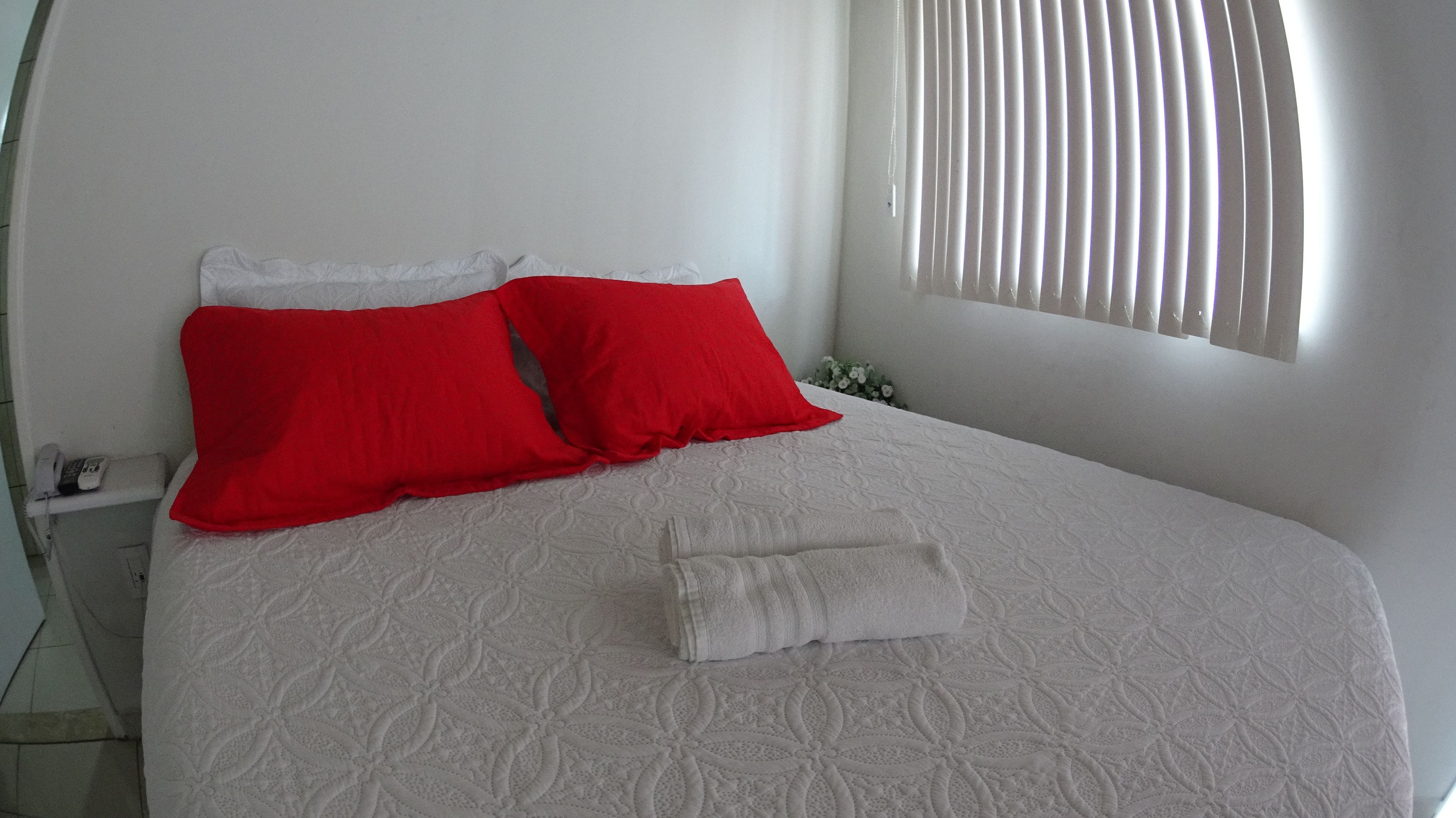 A neatly made bed with a white bedspread and two red pillows, a white bedside table with a phone, and a window with blinds.