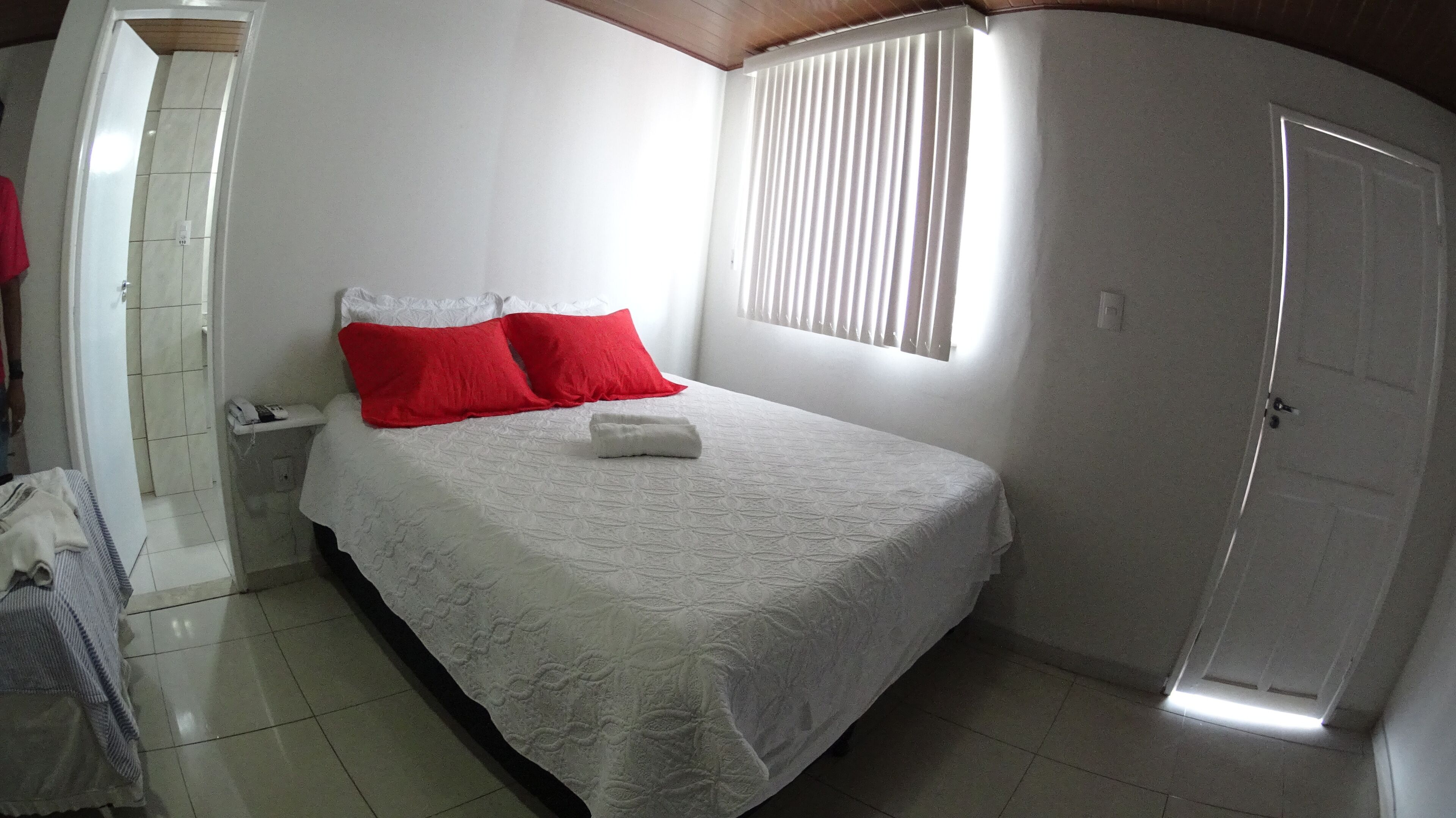 A bedroom with a bed, a red pillow, a white pillow, a white blanket, and a white towel.