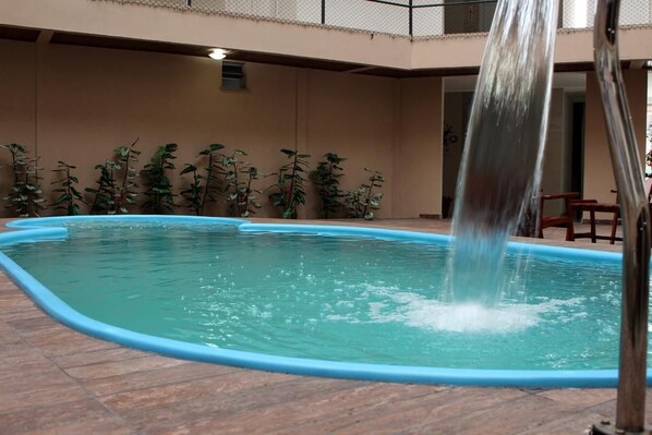 An indoor swimming pool with a blue border, surrounded by artificial plants and a tiled floor.