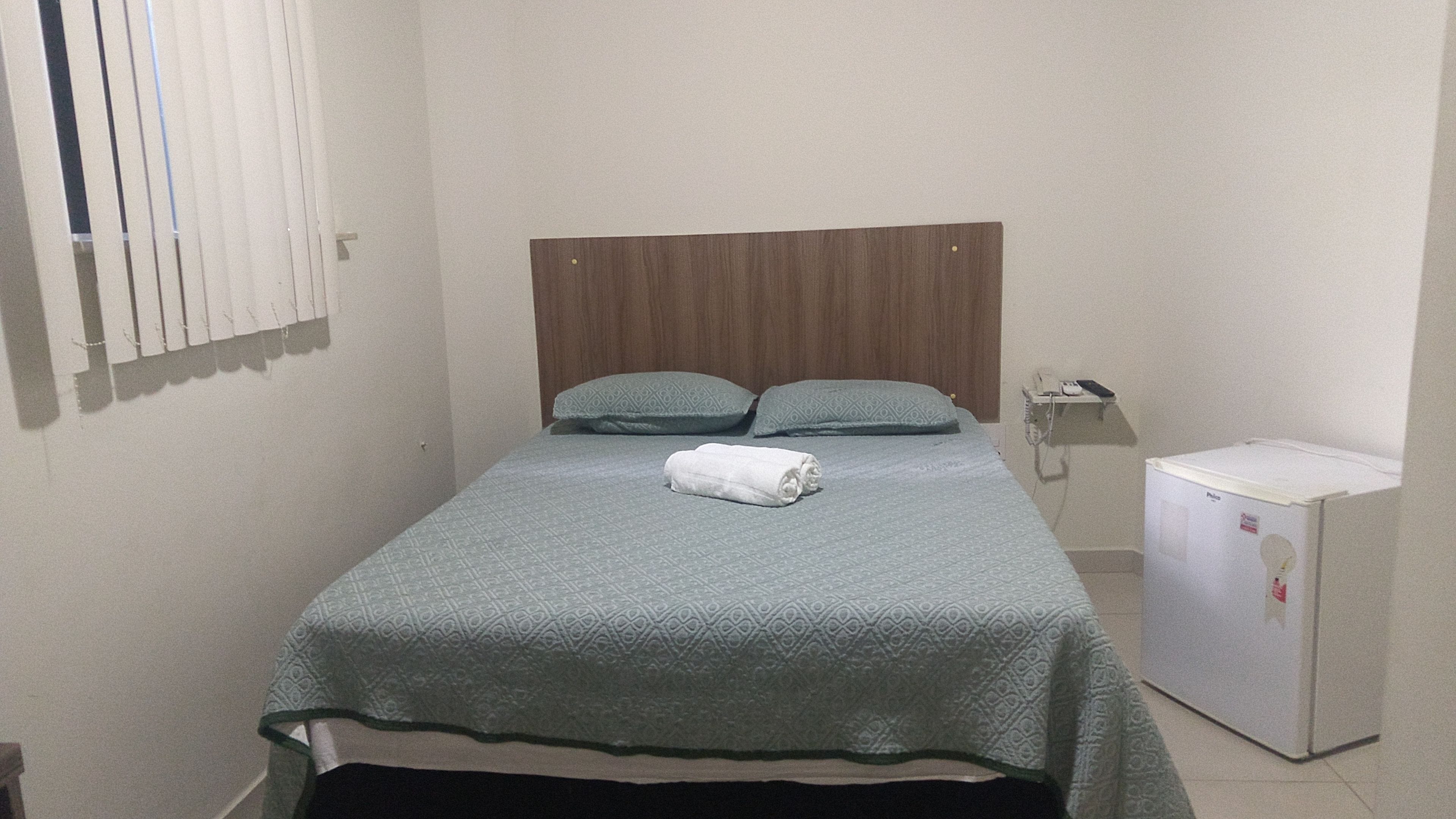 A small, neatly made bed with a patterned bedspread and two pillows, a small white refrigerator, and a bedside telephone.