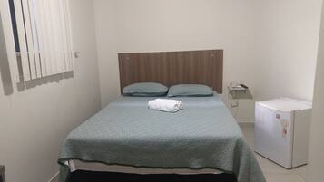 A small, neatly made bed with a patterned bedspread and two pillows, a small white refrigerator, and a bedside telephone.