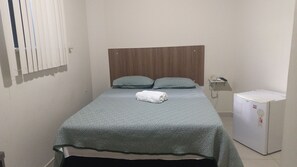 Un petit lit bien fait, avec une couvre-lit à motifs et deux oreillers, un petit réfrigérateur blanc et un téléphone sur la table de chevet.