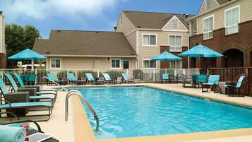 A swimming pool with lounge chairs and umbrellas, surrounded by buildings with a brick facade and a clear blue sky.