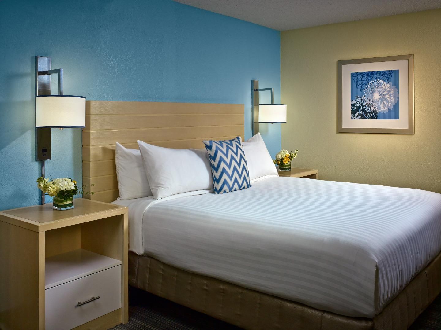 A hotel room with a bed, two bedside tables, a nightstand, a drawer, a wall-mounted lamp, and a framed picture.
