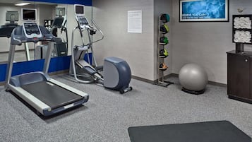 A gym with treadmills, elliptical machines, a stationary bike, a wall-mounted TV, a motivational poster, a rack of helmets, and a large exercise ball.