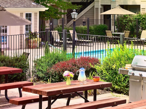 Une terrasse avec des tables et des bancs en bois, un barbecue et une piscine en arrière-plan.