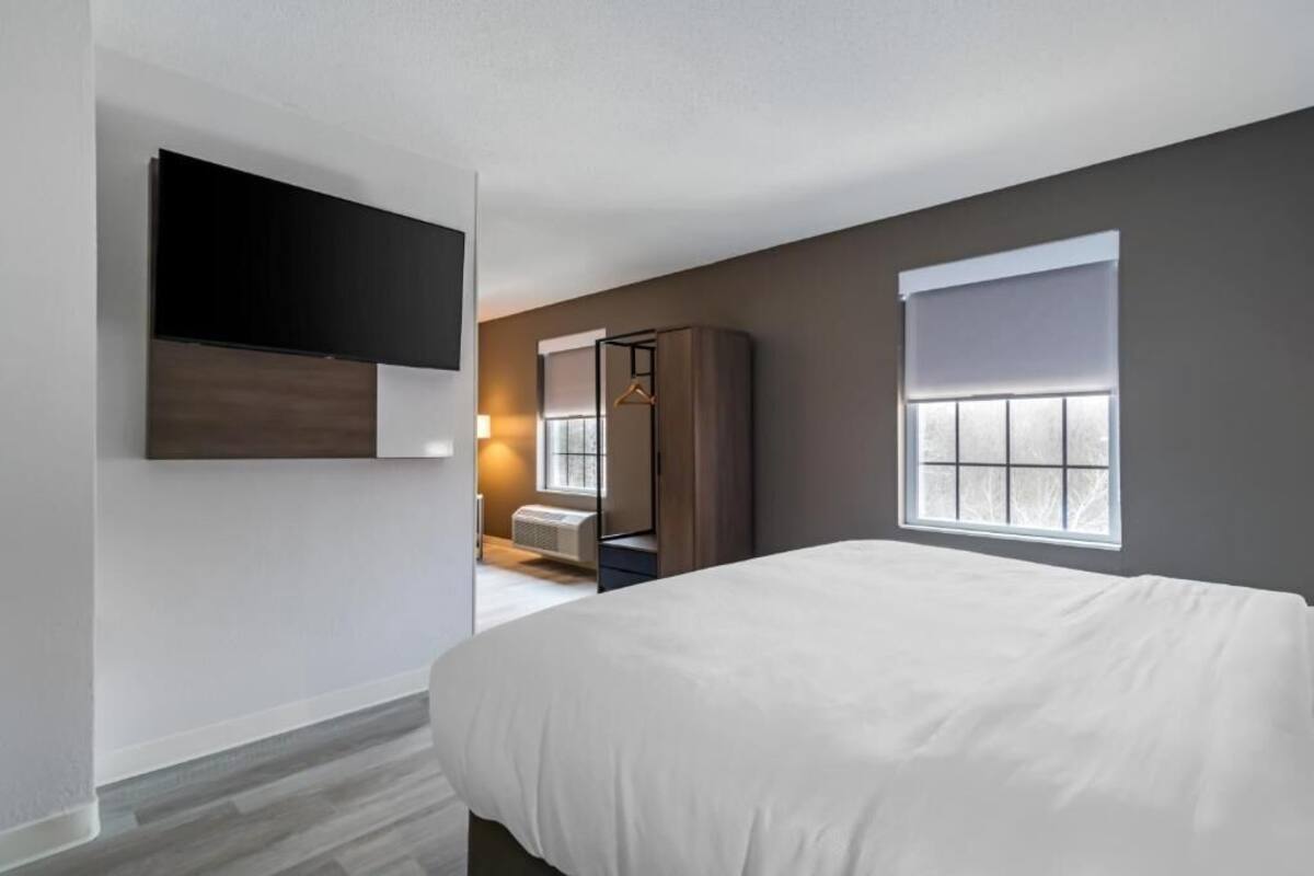 A modern hotel room with a large bed, a flat-screen TV mounted on the wall, a wardrobe, and a window with blinds.