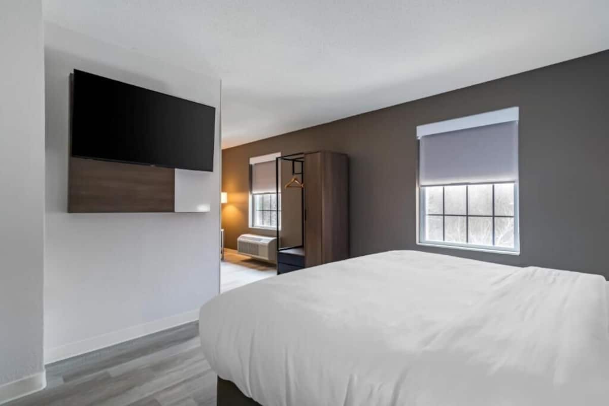 A modern hotel room with a large bed, a flat-screen TV mounted on the wall, a wardrobe, and a window with blinds.