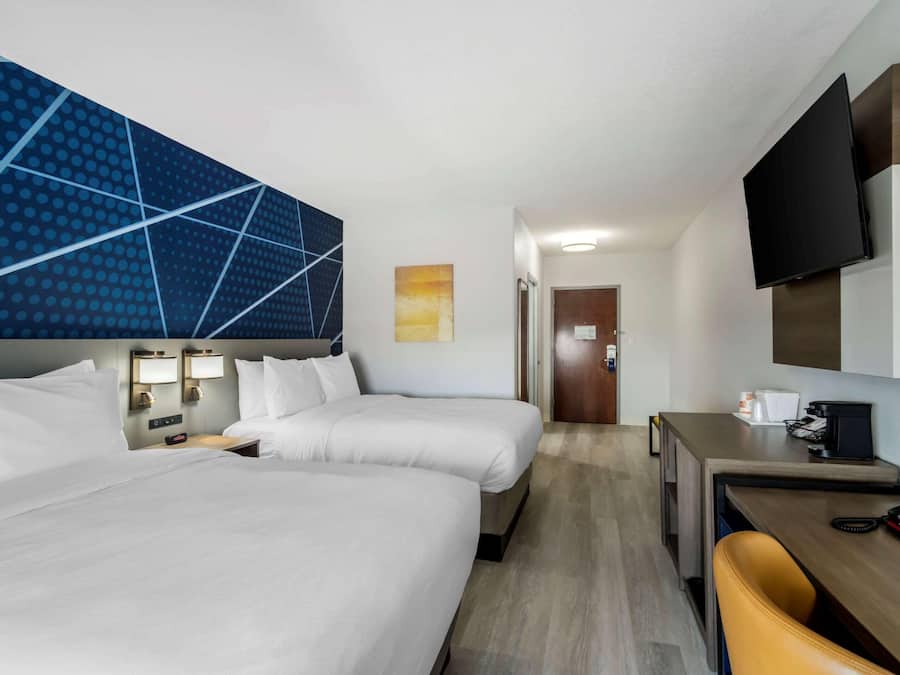 Quarto de hotel com duas camas, uma escrivaninha, uma cadeira, uma televisão e a porta do banheiro.