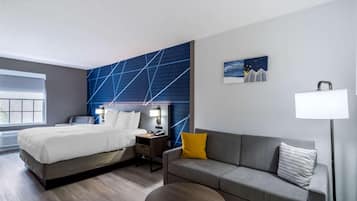A modern hotel room with a bed, a sofa, a small table, a nightstand, a lamp, and a window.