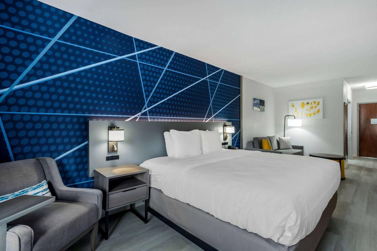 A modern hotel room with a large bed, a desk, a chair, and a wall with a geometric pattern.
