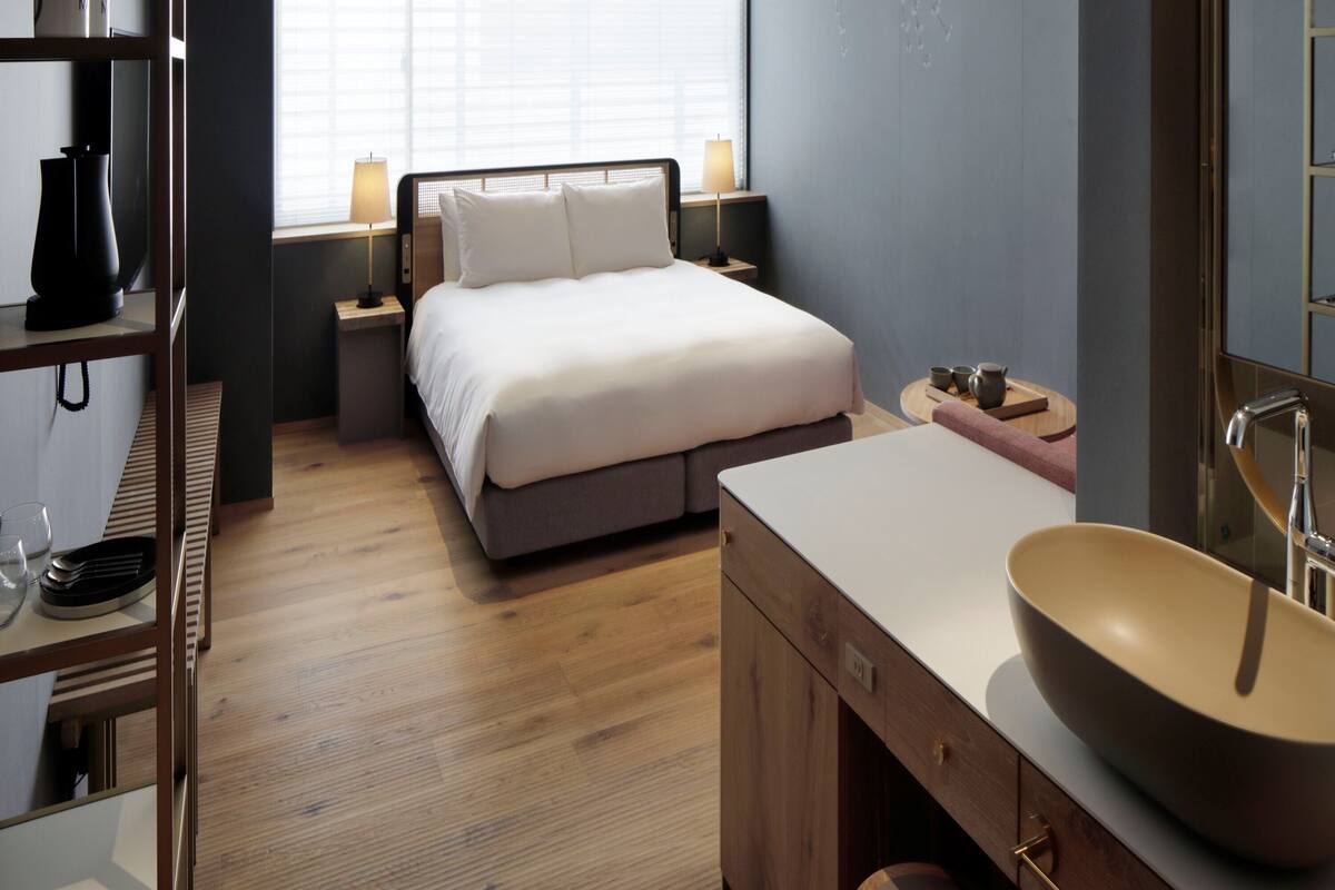 Une chambre moderne avec un grand lit, des tables de chevet, une coiffeuse avec un grand lavabo et une fenêtre avec des stores.