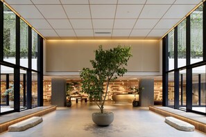 Un vestíbulo amplio con un árbol central, ventanas grandes y una zona de recepción al fondo.