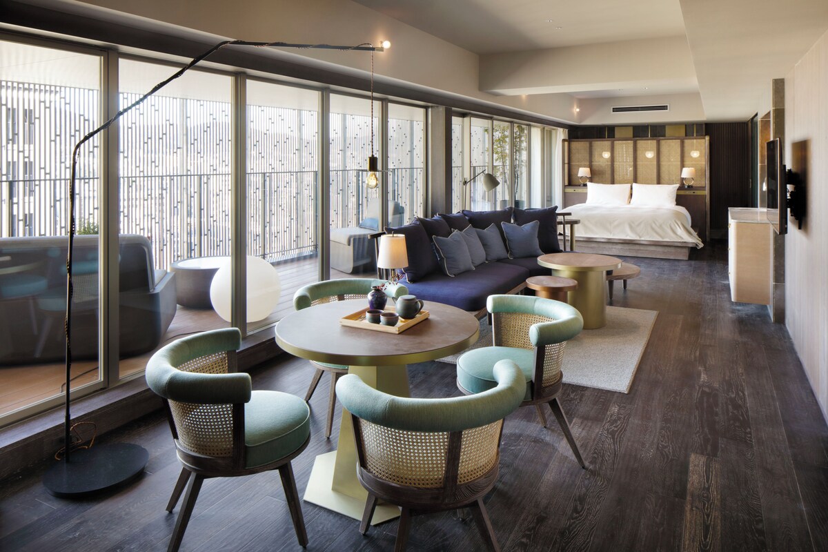 Habitació d'hotel moderna amb un llit gran, un sofà, una taula de menjador amb cadires i vistes per grans finestres.