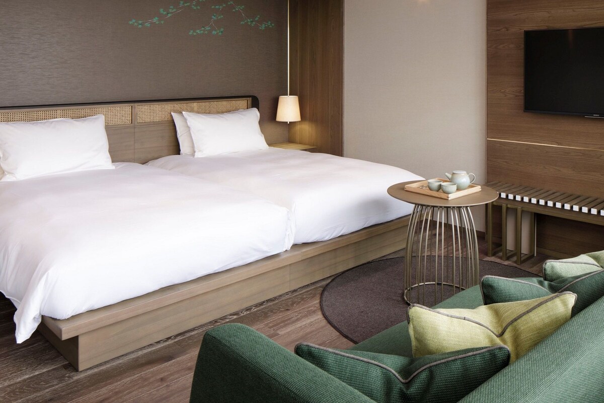 A hotel room with a large bed, a small round table with a tray of tea, a sofa, and a TV mounted on the wall.