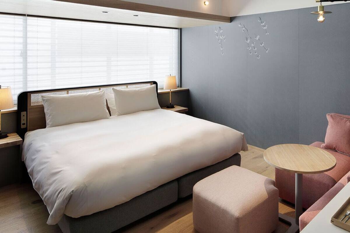 Une chambre d’hôtel moderne avec un grand lit, un canapé, une petite table ronde et une fenêtre avec des stores.