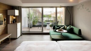 Une chambre d’hôtel moderne avec un lit, un canapé vert, une petite table et un balcon aménagé avec du mobilier d’extérieur.