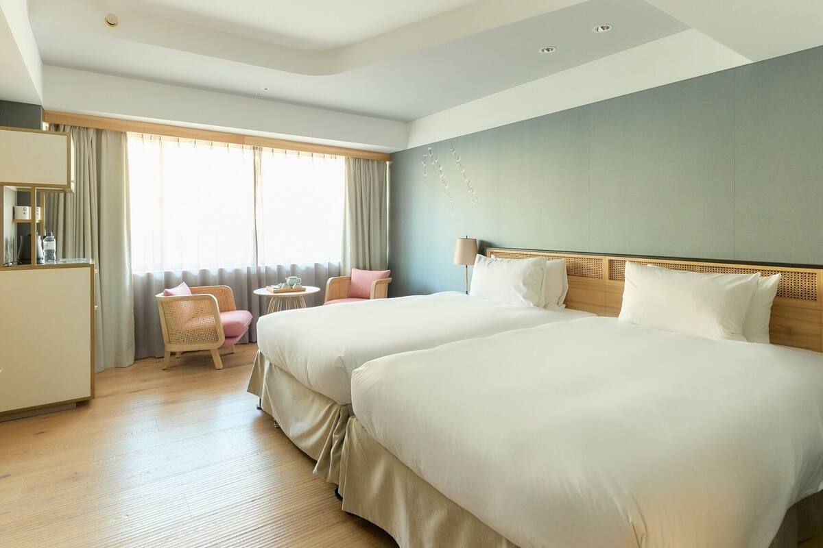 Une chambre d’hôtel avec deux lits, un bureau, une chaise et une fenêtre avec des rideaux.