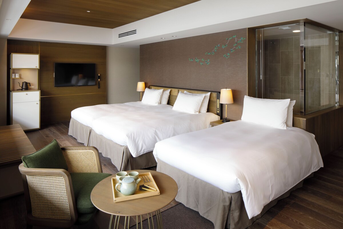 A hotel room with two beds, a TV, a seating area with a table, and a shower area.