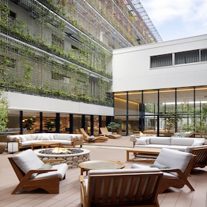 A modern outdoor lounge area with seating and a fire pit, surrounded by a building with a glass facade and greenery on the upper levels. - Good Nature Hotel Kyoto (Kyoto)