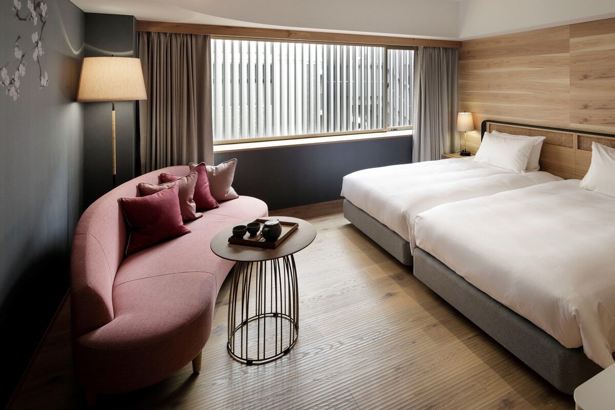 A hotel room with a sofa, two beds, a small table, and a window with blinds.