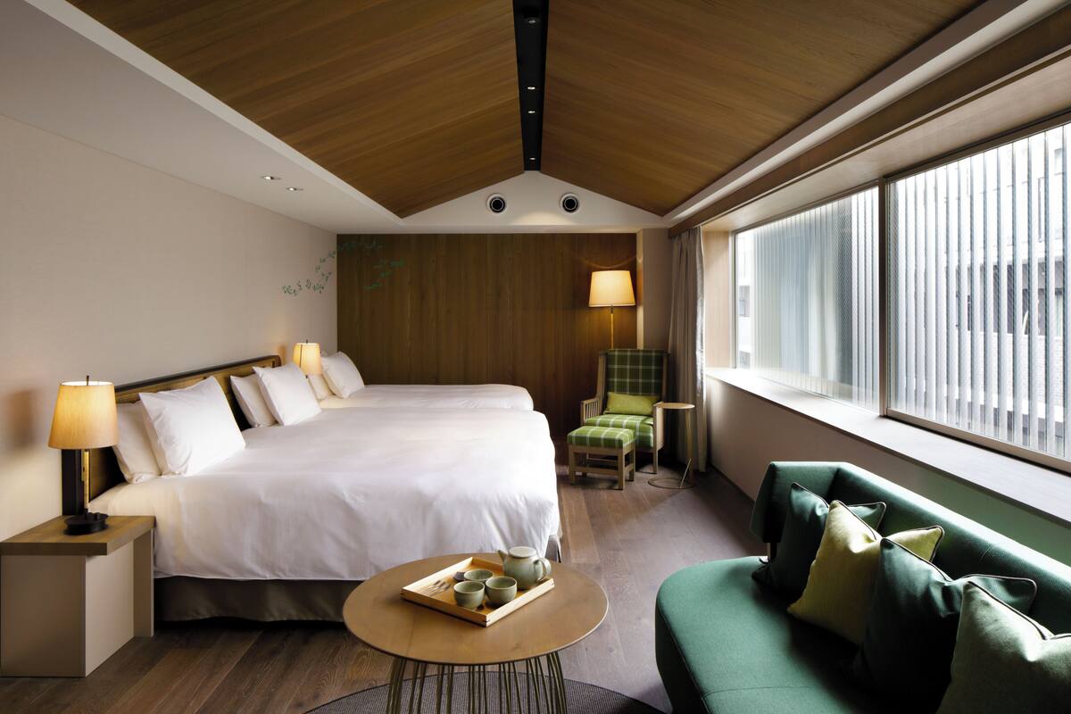 Une chambre d’hôtel avec un grand lit, un canapé vert, une petite table sur laquelle se trouve un plateau avec des tasses à thé et une théière, et une fenêtre avec des stores.