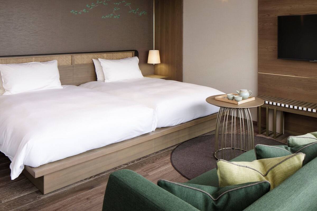 Une chambre d’hôtel comprenant un lit, une télévision, une petite table avec un plateau contenant divers objets, et un coin salon avec des coussins.