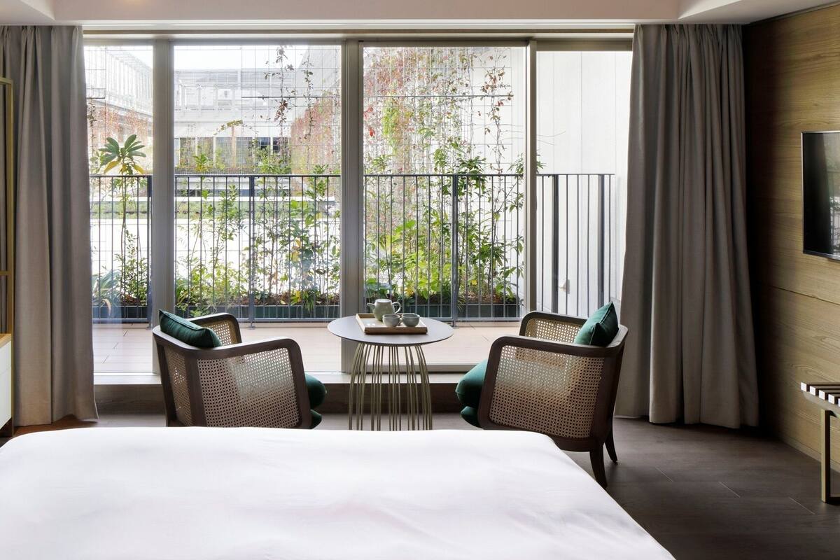 Une chambre d’hôtel avec un grand lit, deux chaises, une petite table, une télévision et un balcon agrémenté de plantes.